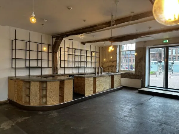 Interieur van een leeg horecapand aan het Sint Agathaplein met een onafgewerkte houten bar, metalen wandrekken en zicht op het plein door grote ramen.